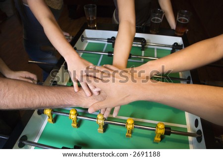 Group of young friends clasping hands in solidarity over foosball table.