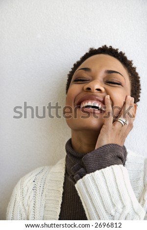 Close up head and shoulder of African-American woman standing against white wall smiling with hand on face and eyes closed.