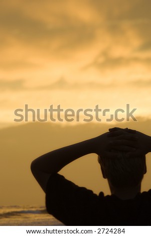 Caucasian pre-teen boy sihouette with hands behind head on beach at sunset.