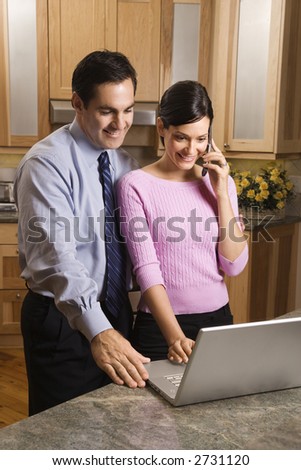 MId-adult female talking on phone and mid-adult male both looking at laptop computer in kitchen.