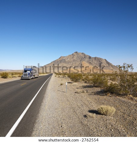Tractor trailer driving on desert road with mountain in background.