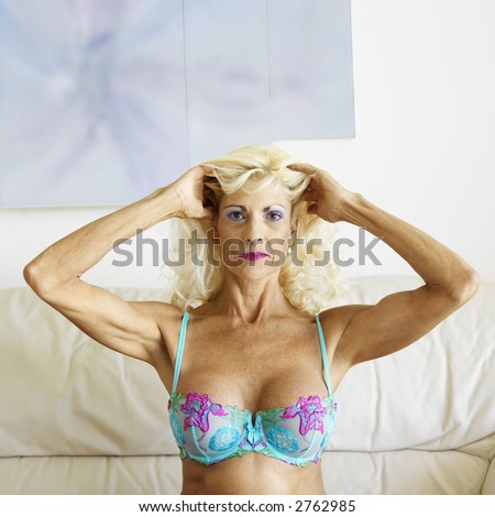 Tan Caucasion blonde middle-aged woman posing in bra with arms over head.