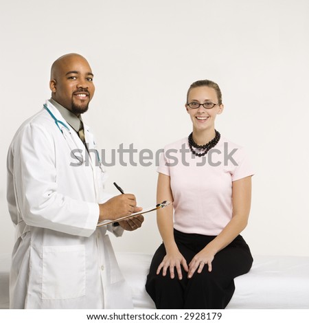 Mid-adult African-American doctor with clipboard and Caucasion mid-adult female patient in doctor\'s office looking at viewer and smiling.