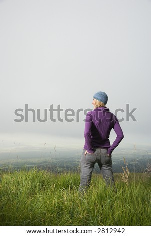 Back view of smiling middle-aged Caucasian woman standing alone in field looking to side.