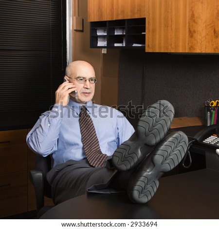 Caucasian middle-aged businessman in office sitting with feet on desk talking on cellphone.