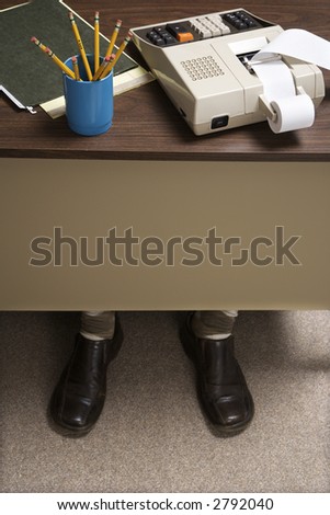 Feet of retro businessman sticking out of bottom of desk with notebook, pencils and adding machine.
