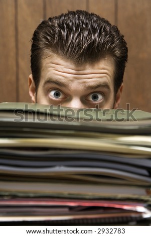 Caucasion mid-adult retro businessman peering with wide eyes over a tall stack of folders.