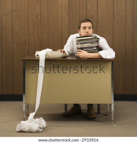 Mid-adult Caucasian male holding stack of paperwork with hand on calculator with long printout.