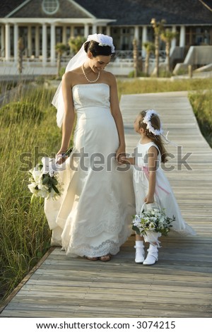 Caucasian mid-adult bride holding hands with flower girl walking down wooden beach walkway.