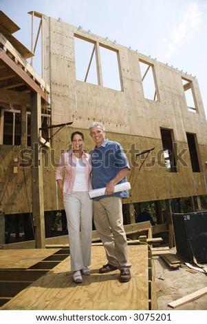 Caucasian mid-adult male holding blue prints with arm around mid-adult female in building construction site.