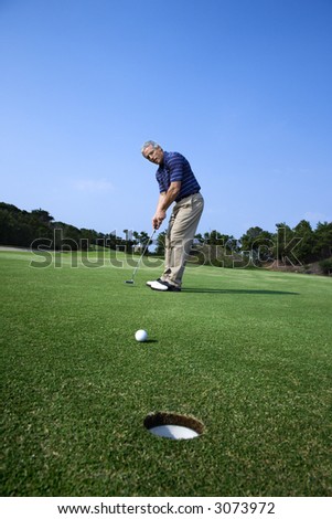 Caucasion mid-adult man putting golf ball with hole in foreground.