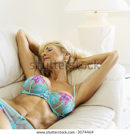 Tan Caucasion blonde middle-aged woman seductively lying in underwear on couch with arms raised.