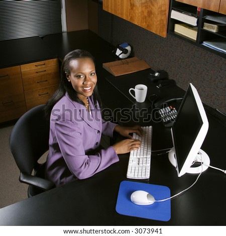 High angle view of African-American young adult business woman working at computer in office smiling and looking at viewer.