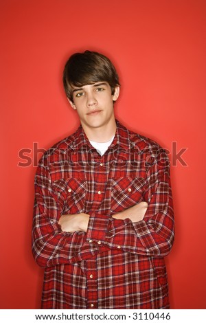 Portrait of Caucasian teen boy with arms crossed standing against red background wearing flannel shirt.