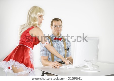 Caucasian young man smiling dressed like nerd sitting at computer looking at viewer with Caucasian young blonde woman in french maid outfit bending over and pointing.