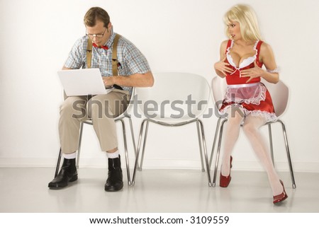 Caucasian young man dressed like nerd sitting with laptop with Caucasian young blonde woman trying to distract him with cleavage.
