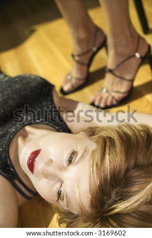 Caucasian mid adult woman lying on wood floor with legs of another woman in background wearing heels.