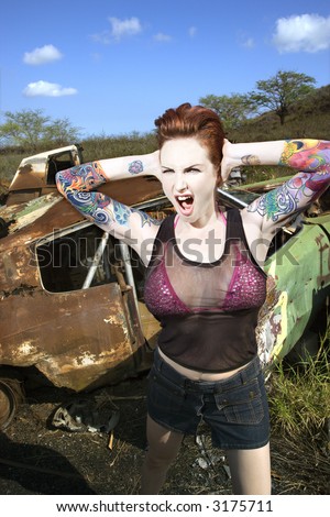 Sexy tattooed Caucasian woman yelling and holding her hands over her ears in junkyard.
