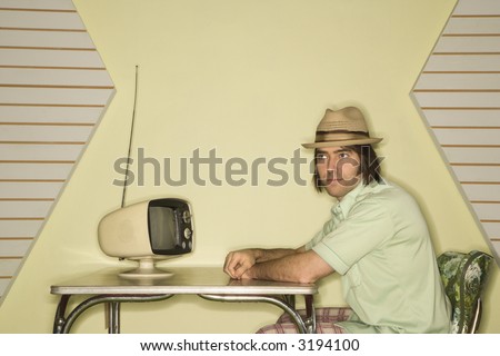Caucasian mid-adult man wearing hat sitting at 50\'s retro dinette set in front of old television.