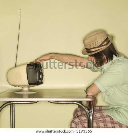 Side view of Caucasian mid-adult man wearing hat sitting at 50\'s retro dinette set tapping old television set.