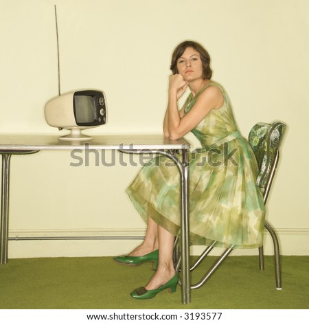 Pretty Caucasian mid-adult woman wearing green vintage dress sitting at 50\'s retro dinette in front of old televsion looking bored.