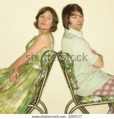 Caucasian mid-adult couple wearing vintage clothing sitting back to back in green vinyl chairs with arms crossed looking angry.