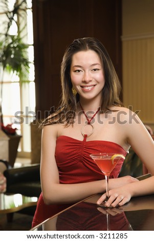 Taiwanese mid adult woman in red dress smiling and standing at bar with drink.