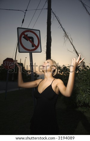 Caucasian mid-adult blonde woman wearing little black dress posing with hands up beside no left turn sign.