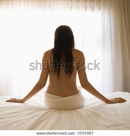 Back view portrait of pretty Caucasian young woman partially nude sitting on bed by sunlit window.