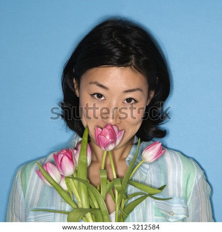 Head and shoulder portrait of pretty Asian mid adult woman smelling flowers on blue background.