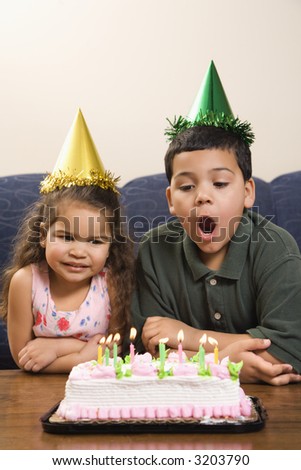 Hispanic girl and boy wearing party hats preparing to blow candles out on birthday cake.