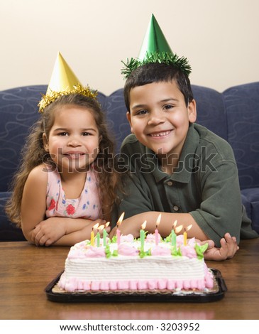 Hispanic girl and boy wearing party hats sitting in front of birthday cake smiling and looking at viewer.