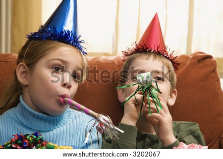 Caucasian children looking bored wearing party hats and blowing noisemakers.