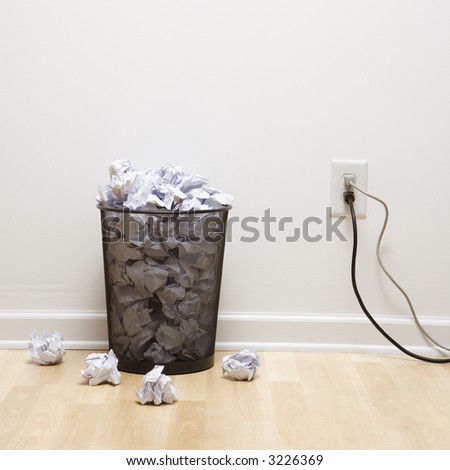 Full wire mesh trash can with crumpled paper next to electrical outlet and plugs.