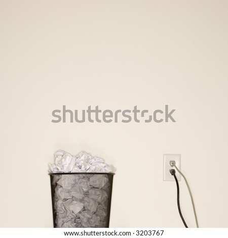 Full wire mesh trash can with crumpled paper next to electrical outlet and plugs.