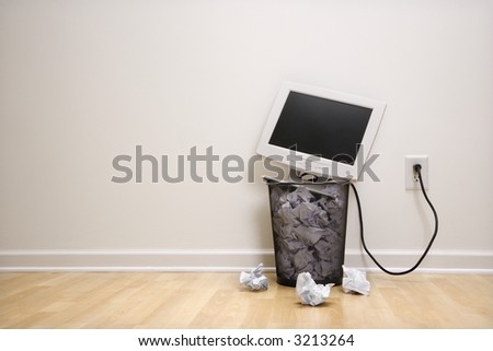 Computer monitor in trash can surrounded by crumpled up paper.