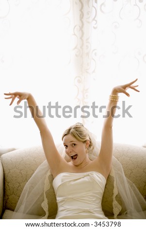 Caucasian bride on love seat with arms raised and joyful expression.