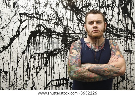 Caucasian tattooed man with braided beard standing against paint splattered background.