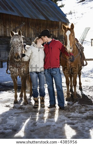 Caucasian couple kissing and holding horses with stable in background.