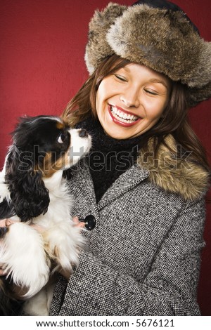 Young adult Caucasian woman wearing fur hat holding King Charles Spaniel in arms and smiling.