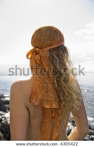 Back view of nude Caucasian mid-adult woman with wavy hair and head scarf at coast.