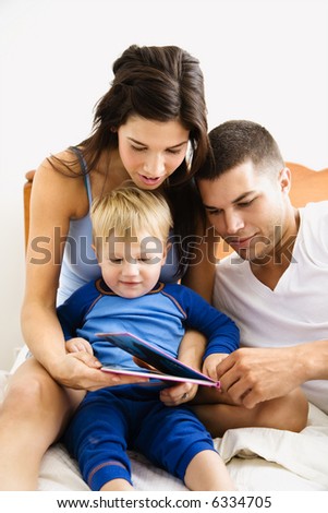 Caucasian parents and toddler son reading book in bed.