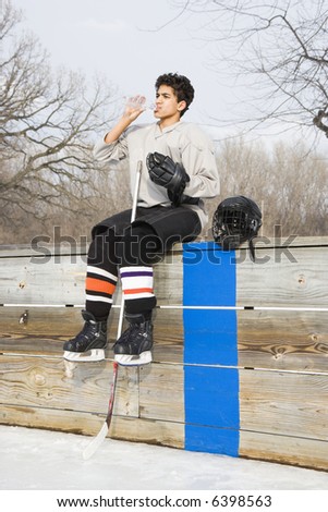 Boy in ice hockey uniform holding hockey stick sitting on sidelines drinking water.