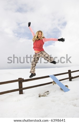Young woman in winter clothes by fence in snowy field with snowboard jumping into air excitedly.
