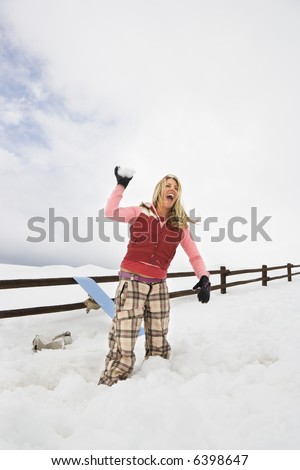 Young woman in winter clothes by fence in snowy field laughing and ready to throw snowball.