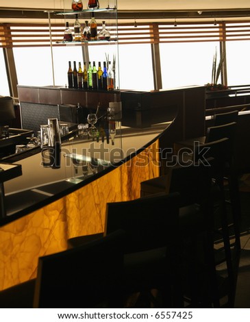 Empty bar in restaurant with row of barstools and windows in background.