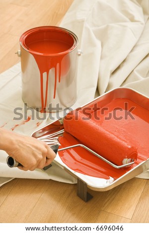 Hand holding paint roller in tray with painting supplies on drop cloth.