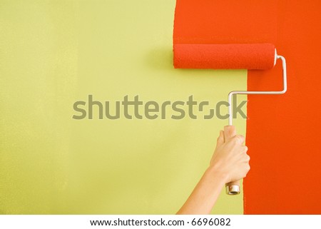 Caucasian female hand painting red over a green wall with a paint roller.