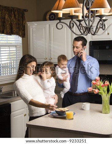 Caucasian mother and father in kitchen busy with children and cellphone.