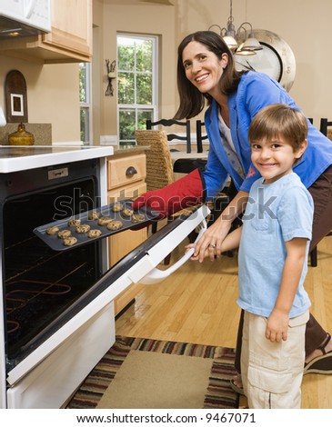 Hispanic mother and son putting cookies into oven and smiling at viewer.